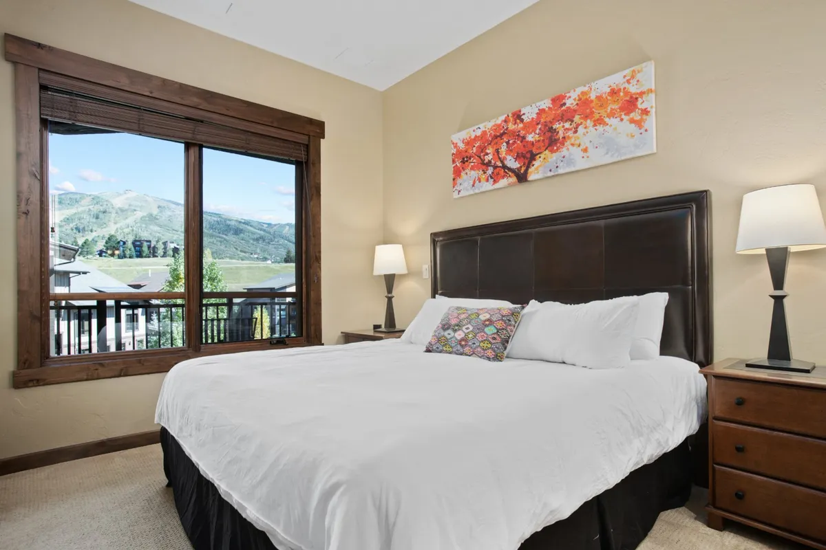 This image features a spacious bedroom with a large window showcasing breathtaking mountain views. The bed is outfitted with plush, inviting bedding and complemented by colorful artwork and soft lighting from nightstand lamps, radiating a tranquil, restful mood.