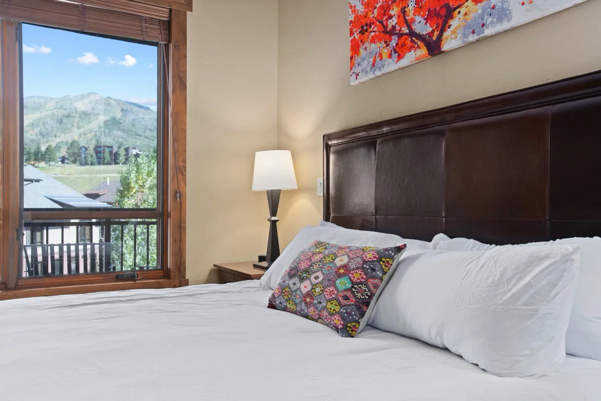 A cozy bedroom space with a comfortable bed dressed in colorful pillows, artistic wall decor, and a window that frames beautiful mountain scenery. The setting invites relaxation and quiet moments of comfort.
