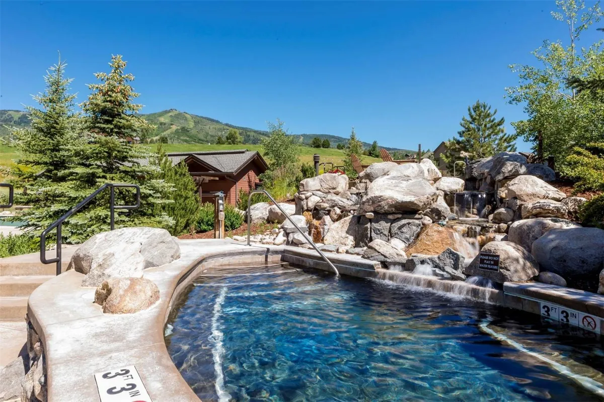 A tranquil hot tub set among natural rock formations and lush greenery, with a cascading waterfall and mountain views in the distance. The setting is serene and luxurious, ideal for unwinding after outdoor adventures.
