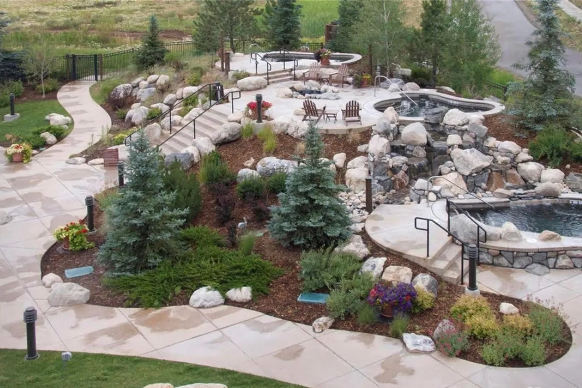 This exterior shot features rock formations, inviting hot tubs, seating areas, and landscaped paths, all bathed in a serene mountain atmosphere. The setting evokes relaxation and social enjoyment after a day of adventure.