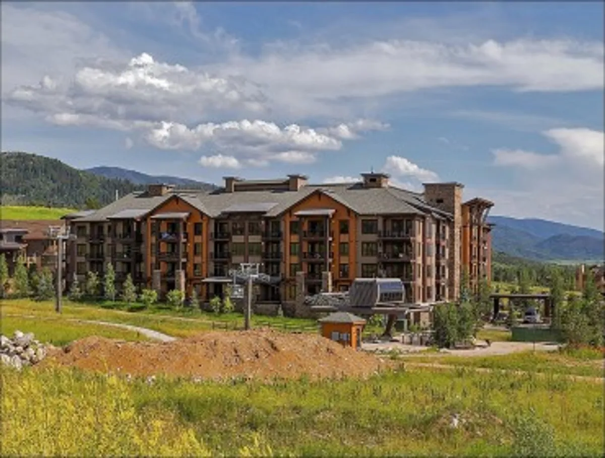 A multi-story lodge building sits against a lush green landscape and a panoramic mountain backdrop, with a ski lift visible nearby. The scene is vibrant and active, with a sense of adventure in both ski season and summer months.