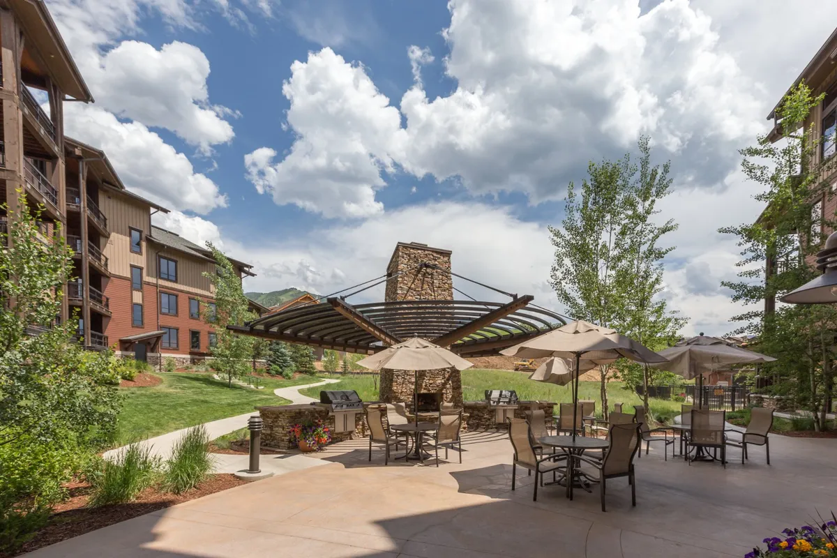 This image shows a well-appointed outdoor seating area featuring comfortable furniture, a fireplace, and lush landscaped gardens under a cloudy sky. The scene suggests spaces perfect for relaxing, social gatherings, and enjoying fresh air with mountain vibes, while the grill in the background hints at opportunities for outdoor dining.