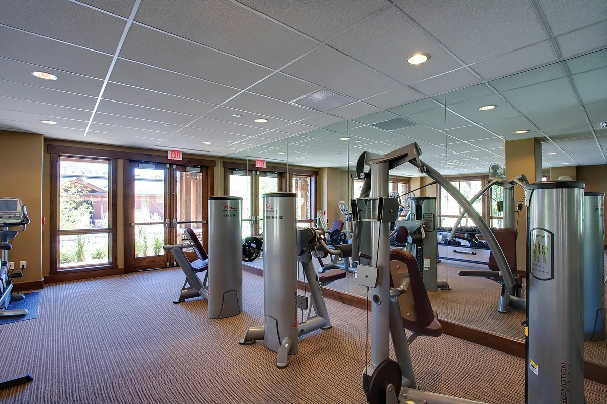 The image features a bright fitness center with a row of exercise machines facing large windows that let in natural light and overlook greenery. The mirror wall enhances the sense of spaciousness and motivates healthy routines during extended stays.