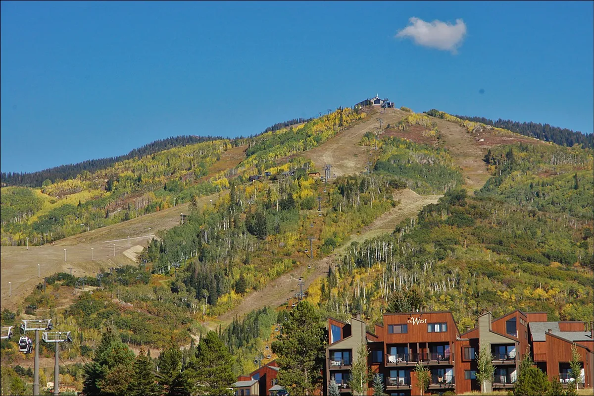 A dynamic view highlights the ski resort on the mountain, colorful trees, and modern buildings in the foreground, with the ski lift prominently visible. The setting conveys both the proximity to outdoor activities and the comfort of nearby accommodations.