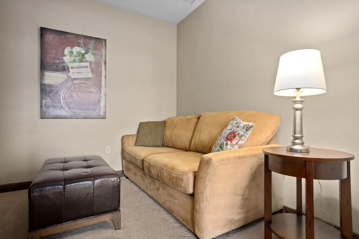A flexible den area featuring a comfortable sofa, framed artwork, a well-placed lamp, and an ottoman. The space feels intimate and is ideal for reading, working, or providing extra sleeping arrangements.