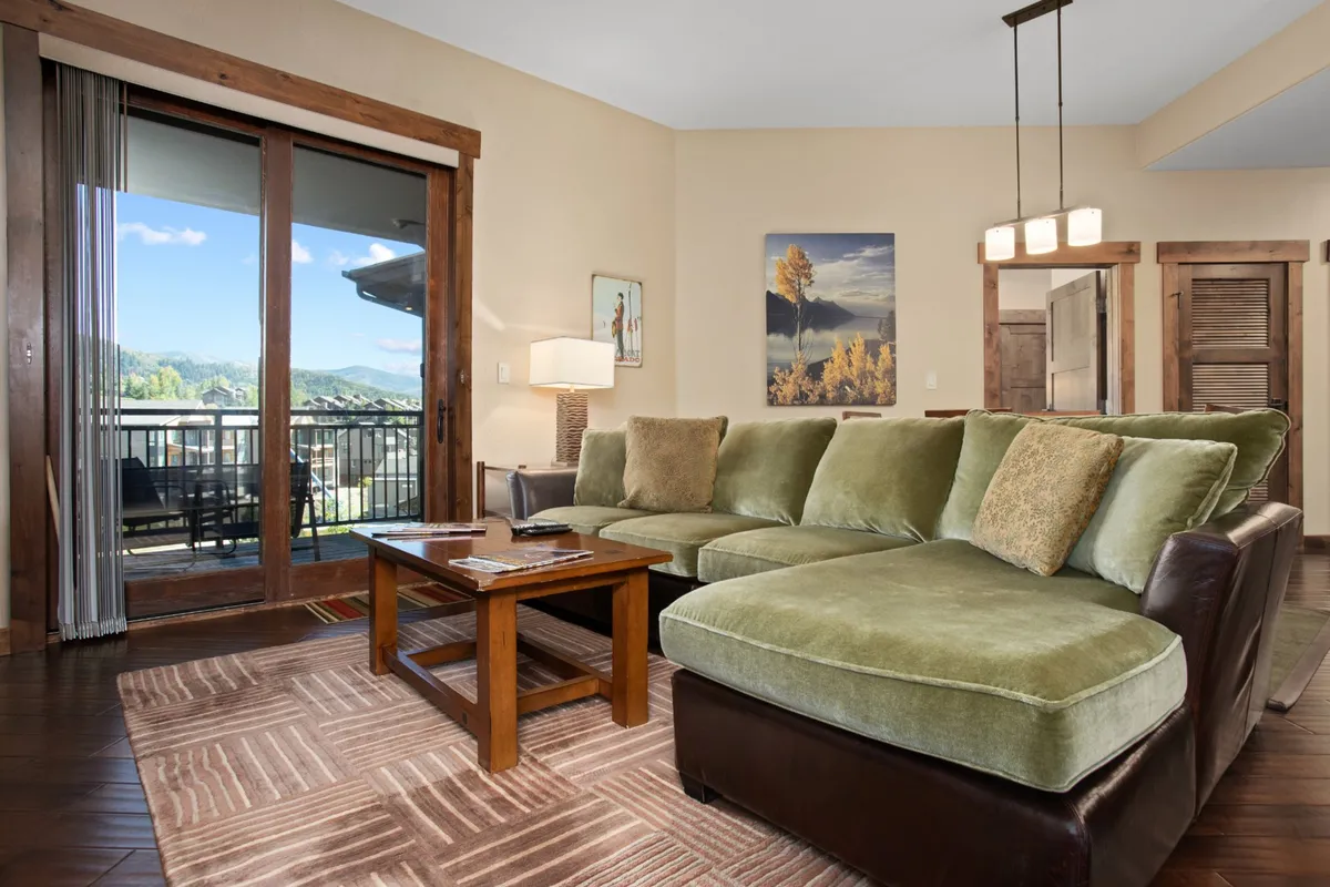 A spacious living area featuring a green sectional sofa and a wooden coffee table, set against large windows framing expansive mountain views. Artwork adorns the wall, and the room is bathed in natural light, creating a cozy and welcoming atmosphere ideal for relaxing or socializing.
