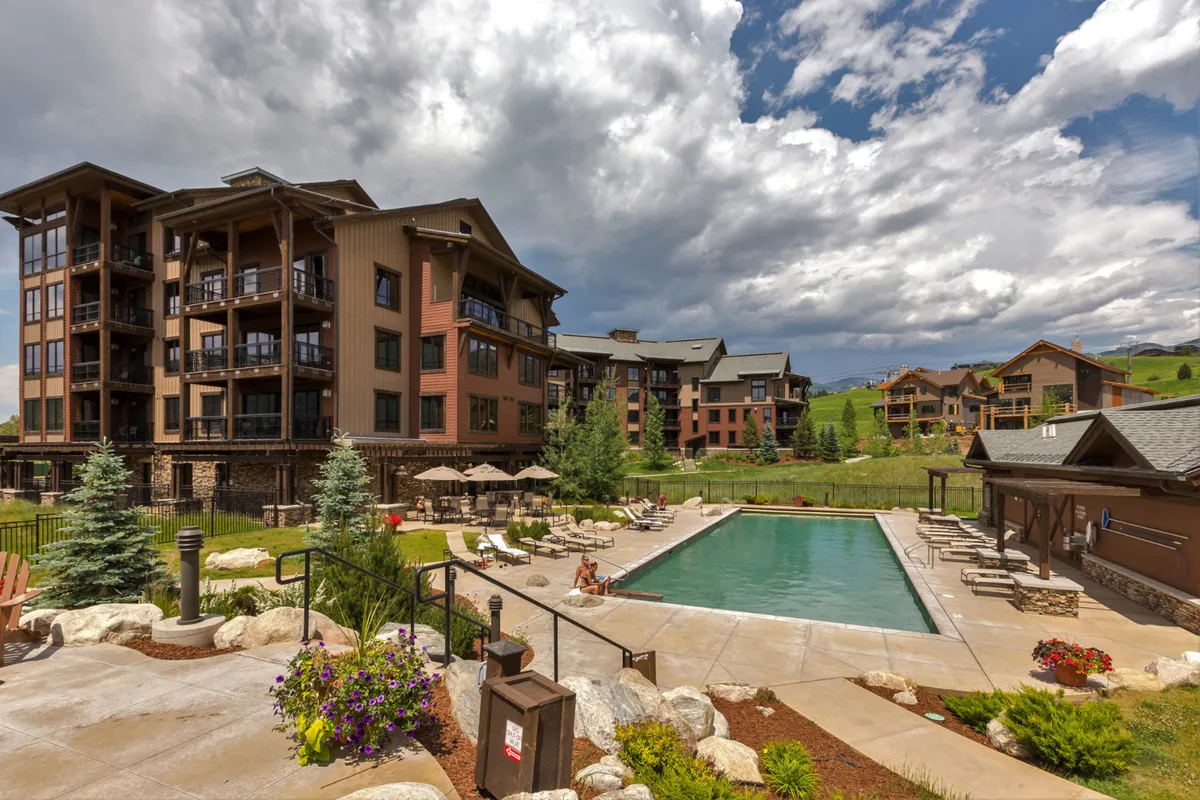 A tranquil scene of a large heated outdoor swimming pool surrounded by lounge chairs, landscaped gardens, and neighboring buildings, perfect for social gatherings or a peaceful dip after a day on the slopes.