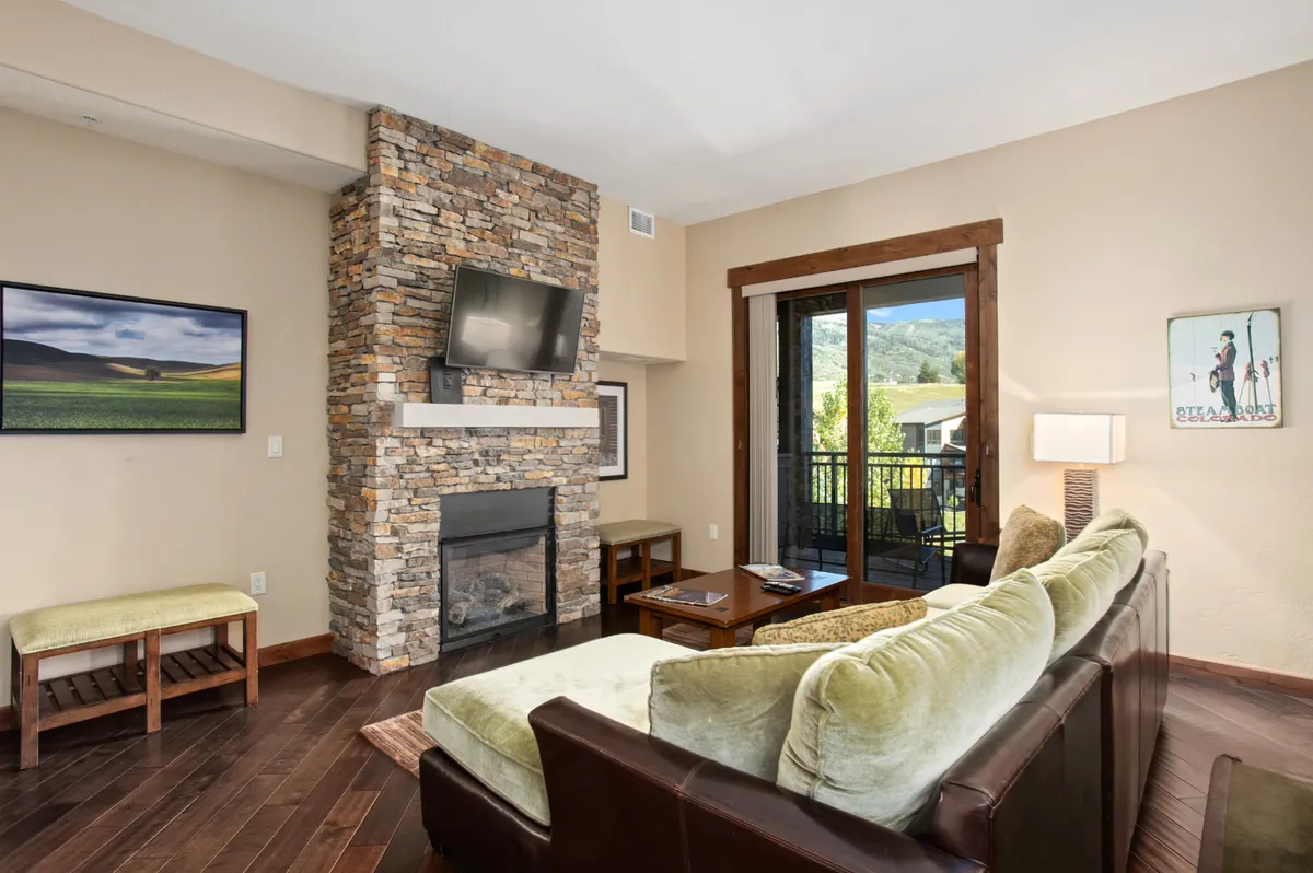 This inviting interior image showcases a spacious living area with a stone fireplace, large windows offering a view of the outdoors, comfortable seating, and a TV mounted on the wall. The arrangement encourages relaxation, socializing, and enjoying the scenic views from inside, blending indoor comfort with outdoor inspiration.